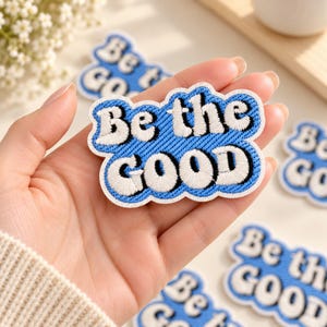 May include: An embroidered patch with a blue background and white text that reads Be the GOOD. The patch has a scalloped white border and a black outline around the letters. The text is arranged in two lines with a bold, rounded font style.