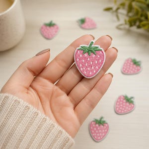 May include: A small, heart-shaped strawberry embroidered patch featuring a pink body with white seed details and a green stem. The patch has a white border and is designed for fabric application, showcasing detailed thread work and vibrant colors.