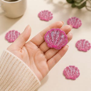 May include: A collection of small, pink seashell-shaped patches decorated with shimmering sequins. Each patch features a fan-like design with distinct ridges and a bright pink stitched border, creating a textured and reflective appearance.