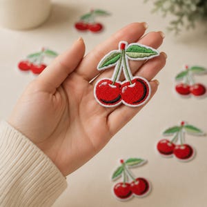 May include: An embroidered cherry patch featuring two bright red cherries with dark red shading, two green leaves, and thin green stems. The patch has a white border and is shown against a neutral background with several identical cherry patches.