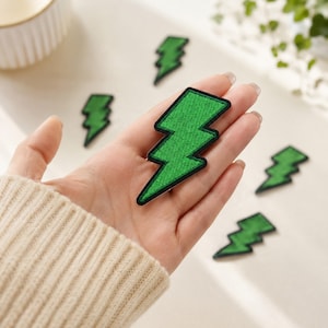 May include: A collection of green lightning bolt patches with black embroidered borders. One patch is held in the palm of a hand, while four additional identical lightning bolt patches are arranged on the surface nearby for comparison.