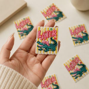 May include: A rectangular embroidered patch featuring a green alligator wearing a pink bow, set against a background of vertical yellow and white stripes. The word LATER is stitched in pink letters at the top, accompanied by small star accents.