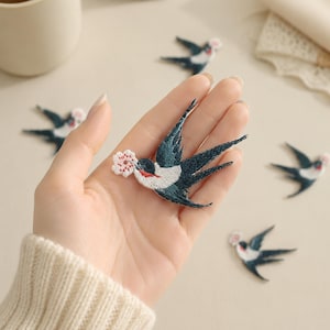 May include: An embroidered swallow patch featuring dark blue wings, a white chest, a red throat accent, and a small pink flower held in its beak. The swallow is depicted in a flying pose with wings extended and a forked tail, showcasing detailed stitching.