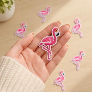 May include: A collection of small, embroidered flamingo patches featuring vibrant pink thread, white accents, and a black beak detail. Each patch depicts a flamingo standing on one leg, with one patch held in the palm of a hand and others scattered.