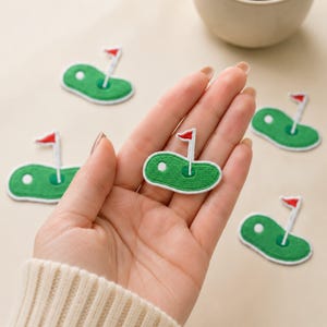 May include: Several embroidered iron-on patches shaped like golf greens. Each patch features a green oval base with a white golf ball, a white flagstick, and a small red flag at the top. The patches are rendered in a flat, stitched design.