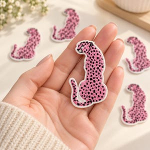 May include: A collection of embroidered iron-on patches, each shaped like a sitting leopard. The patches feature a vibrant pink base color with detailed black spots and a white border, showcasing a consistent feline silhouette design.