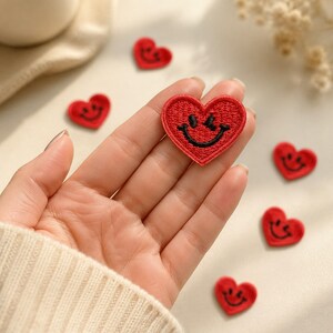 May include: A collection of small, red heart-shaped embroidered patches, each featuring a black stitched smiley face. One patch is held in the palm of a hand, while several other identical patches are scattered around it on a light-colored surface.