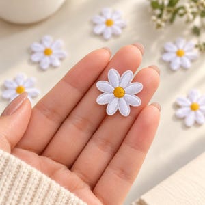 May include: A small, embroidered daisy patch featuring white petals and a bright yellow center. The flower shape is circular with eight distinct petals. Several identical daisy patches are scattered around the central piece.