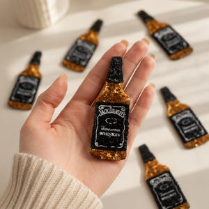 May include: A sequined patch shaped like a whiskey bottle with a black neck and base, featuring a black label with white text that reads Jack Daniel's Tennessee Whiskey. The patch is embellished with gold and black sequins throughout the design.