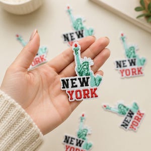 May include: An embroidered patch featuring the Statue of Liberty in green with a red torch flame. Below the statue, the text NEW YORK is stitched in black and red thread. The patch has a white border and is designed for fabric application.