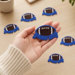 May include: A brown and white football-shaped patch with a textured surface and blue embroidered border. The patch features the text "game day" in blue script lettering at the bottom. Several identical patches are scattered around the main one.