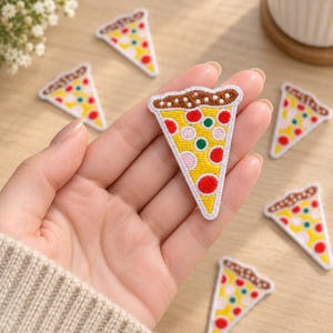 May include: A triangular pizza slice patch featuring a brown crust edge, a yellow cheese base, and embroidered red, green, and light pink circular toppings. The patch has a white border and is shown alongside several identical pizza slice patches.