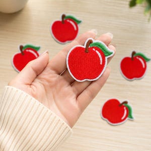 May include: A collection of embroidered apple patches featuring a bright red fruit body, a brown stem, and a single green leaf. Each patch has a white border and a stitched texture, showcasing a consistent design of a round apple with a highlight.