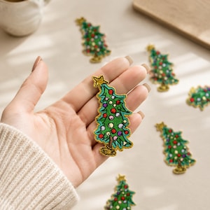May include: An embroidered Christmas tree patch featuring a green tree with colorful circular ornaments and a gold star at the top. The patch has a gold-colored border and is shown in a hand, with several identical patches scattered nearby.