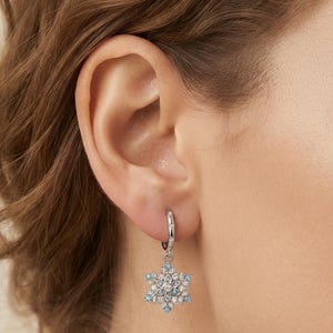 May include: A silver-toned hoop earring featuring a dangling snowflake-shaped charm. The charm is encrusted with small, clear white stones and light blue stones arranged in a symmetrical, six-pointed star pattern, suspended from a small hoop.