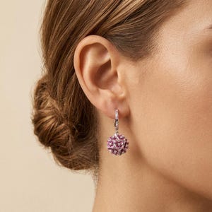 Peut inclure: Une boucle d'oreille créole en argent avec une breloque sphérique pendante composée de multiples petits pétales de fleurs émaillés roses et blancs. L'anneau est orné de minuscules pierres transparentes incrustées, formant un bijou détaillé.