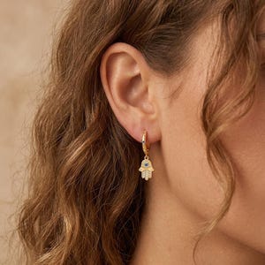 Op de afbeelding: Een gouden oorring met een bungelende Hamsa-handbedel. De bedel is versierd met kleine heldere steentjes en een centrale blauwe steen. De ring zelf bevat ook een klein blauw steendetail, wat zorgt voor een samenhangend en gedetailleerd sieraad.