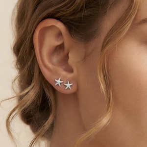 May include: A pair of silver-toned stud earrings shaped like five-pointed starfish. Each earring features a textured surface with a metallic finish, positioned side-by-side on the earlobe to create a cohesive nautical-inspired jewelry set.