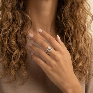 May include: A silver-toned bypass ring featuring two cross-shaped terminals. One cross is plain polished metal, while the other cross is encrusted with small, clear, sparkling stones. The ring has an open, adjustable band design.