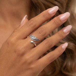 Könnte beinhalten: Ein silberfarbener offener Ring mit einem detaillierten Federmotiv, das sich teilweise um den Finger legt. Das Metall weist in den vertieften Bereichen ein oxidiertes Finish auf, um die Textur der Federadern und die geschwungene Spitze zu betonen.
