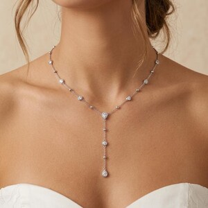 Op de afbeelding: Een zilverkleurige lariat-ketting met een fijne ketting versierd met gelijkmatig verdeelde, ronde, in zetting geplaatste heldere stenen. Het midden van de ketting toont een peervormige steen, met een verticale hangende ketting eindigend in een bijpassende peervormige steen.