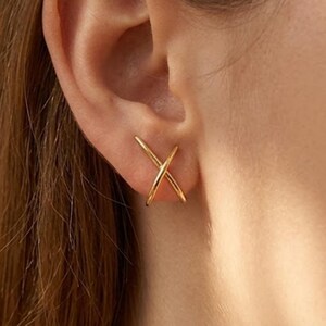 May include: A gold-toned ear cuff featuring a simple, intersecting X-shaped design. The piece consists of two thin, smooth metal bars that cross over each other, resting securely against the outer edge of the ear cartilage without a piercing.