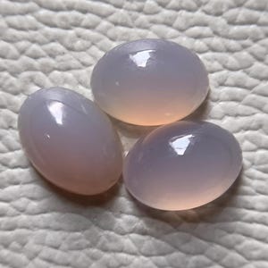 Op de afbeelding: Drie ovaalvormige, gladde, in cabochon geslepen edelstenen met een doorschijnende, licht lavendelgrijze kleur. Elke steen heeft een gepolijst, afgerond oppervlak dat licht reflecteert, wat zorgt voor een zachte, glanzende glans op de stenen.