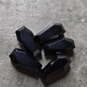 May include: A collection of five small, faceted black beads shaped like miniature coffins. Each bead features a smooth, polished surface with sharp, geometric edges and a pointed top, arranged in a cluster to display their dark, uniform colour.