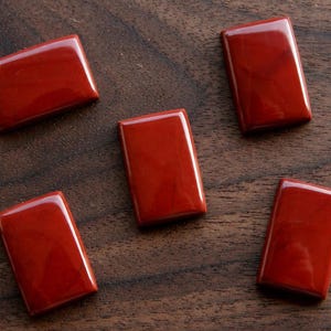 May include: Five polished, rectangular red jasper cabochons with smooth, rounded edges and a glossy finish. The stones are arranged in a scattered pattern, each displaying a deep, solid red color with subtle variations in tone across the surface.