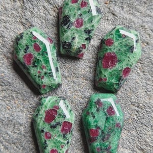 May include: Five polished coffin-shaped gemstones featuring a vibrant green zoisite matrix speckled with deep red ruby inclusions and dark black mineral deposits. Each stone has a smooth, reflective surface with distinct angular facets.