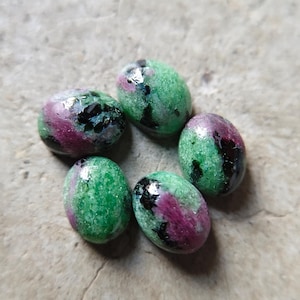 Op de afbeelding: Vijf ovaalvormige cabochon edelstenen met een gepolijst oppervlak, bestaande uit een mix van levendig groen zoisiet, diep magenta robijninsluitingen en donkere zwarte hoornblende mineraalvlekken in een cirkelvormig patroon.