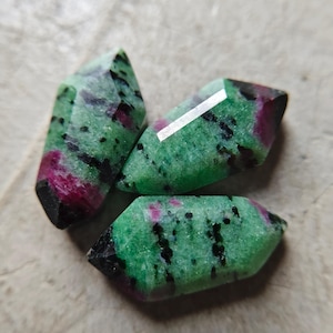 May include: Three faceted gemstones with a pointed marquise shape, featuring a mottled pattern of vibrant green zoisite, deep magenta ruby inclusions, and scattered black hornblende specks throughout the polished, crystalline surfaces.