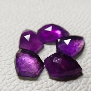 May include: Five faceted, deep purple amethyst gemstones arranged in a circular pattern. Each stone features a polished, multi-faceted surface that reflects light, showcasing a vibrant violet hue and a translucent, crystalline structure.