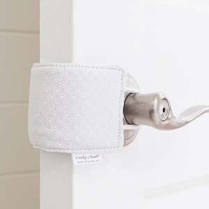 May include: White door stop with a white and gray patterned fabric cover. The door stop is attached to a white door with a silver door handle. The door stop has a white label with the text "Cushy Closer" and the website address "www.cushycloser.com".