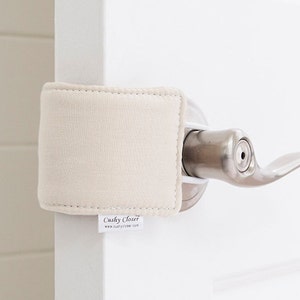 May include: A beige door handle cover with a square design, protecting the door handle. The cover is made of a soft fabric and is attached to the door handle. The tag reads "Cushy Closer" and "www.cushycloser.com".
