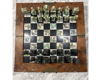 Può includere: Un set di scacchi pieghevole in legno con una scacchiera composta da quadrati neri alternati a quadrati decorati con illustrazioni dettagliate. Il set include pezzi intagliati in verde chiaro e grigio scuro, con una cornice in legno finemente intagliata.