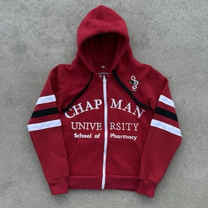 May include: A red zip-up hoodie featuring white text that reads Chapman University School of Pharmacy. The sleeves have white and black horizontal stripes. A small embroidered logo of a snake wrapped around a glass is on the upper chest.