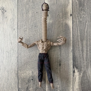 May include: An action figure with a tan, textured, muscular torso and long, thin arms ending in clawed hands. The figure features a long, flexible, accordion-style neck topped with a detailed head and dark hair, wearing black pants with red stitching.