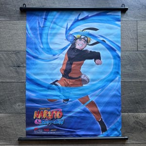 May include: A vertical wall scroll featuring Naruto Uzumaki in an orange and black outfit, surrounded by a swirling blue energy effect. The Naruto Shippuden logo is printed in the bottom left corner, with copyright text below it on a fabric banner.