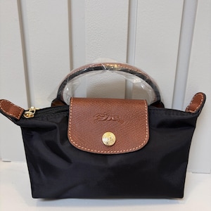 May include: A black nylon handbag featuring a brown leather flap with a gold-tone snap button closure. The bag has a top handle wrapped in protective plastic, a zipper closure, and a debossed horse and rider logo on the front leather flap.
