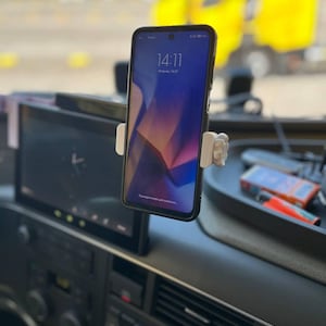 以下が含まれることがあります： 車両のダッシュボードに取り付けられた白いプラスチック製のスマートフォンマウント。14:11と表示された黒いスマートフォンを保持しています。マウントには側面に調整ノブがあり、別のデジタルディスプレイ画面の近くに配置されています。