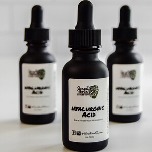 Puede incluir: Tres botellas de vidrio negro de s&eacute;rum facial con cuentagotas. Las etiquetas dicen "Hyaluronic Acid Face Serum with B3 & Caffeine". Las botellas son de Sensational Showers Luxury Handcraft.