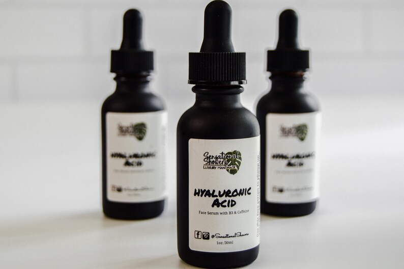 Puede incluir: Tres botellas de vidrio negro de s&eacute;rum facial con cuentagotas. Las etiquetas dicen "Hyaluronic Acid Face Serum with B3 & Caffeine". Las botellas son de Sensational Showers Luxury Handcraft.