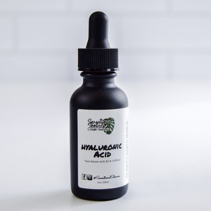 Puede incluir: Una botella de vidrio negro con un cuentagotas, etiquetada como "HYALURONIC ACID Face serum with B3 & Caffeine". La botella tiene un acabado mate y una tapa cuentagotas negra. La etiqueta incluye la marca "Sensational Shores Luxury Handmade".