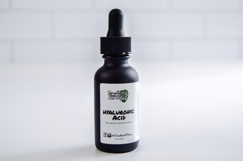 Puede incluir: Una botella de vidrio negro con un cuentagotas, etiquetada como "HYALURONIC ACID Face serum with B3 & Caffeine". La botella tiene un acabado mate y una tapa cuentagotas negra. La etiqueta incluye la marca "Sensational Shores Luxury Handmade".