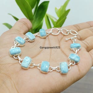 Peut inclure: Un bracelet à maillons en argent composé de neuf pierres précieuses Larimar rectangulaires bleu clair serties dans des cadres en métal poli. Le bracelet comprend un fermoir à bascule et porte le texte Gangajewels925 au centre.