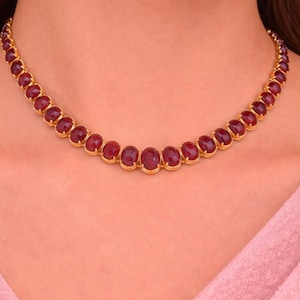Op de afbeelding: Een goudkleurige ketting met een doorlopende rij ovaalvormige, dieprode cabochon edelstenen. Elke steen zit stevig vast in een klauwzetting, wat zorgt voor een uniform, geschakeld ontwerp dat als een klassieke collier rond de hals rust.