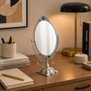 May include: An oval tabletop mirror with an ornate silver-toned frame featuring intricate scrollwork, small dangling charms, and two embedded green oval stones at the top and bottom. The mirror is supported by a decorative pedestal base.
