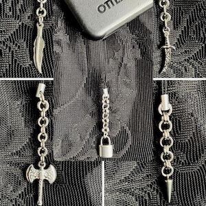 May include: A collection of five silver-toned metal charms attached to black cords, featuring a feather, a dagger, a double-headed axe, a padlock, and a spike. The charms are displayed against a black lace fabric background with a partial OtterBox logo.