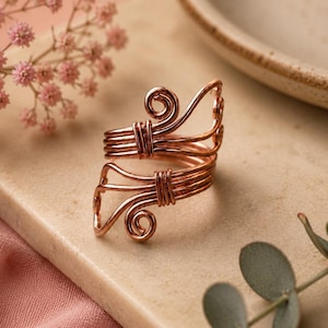 Könnte beinhalten: Ein Ring aus kupferfarbenem Draht mit einem Bypass-Design und zwei offenen Enden. Jedes Ende des Rings ist mit einer dekorativen Spirale versehen und durch kleinere Drahtwicklungen gesichert, die die mehreren Stränge des Rings zusammenhalten.